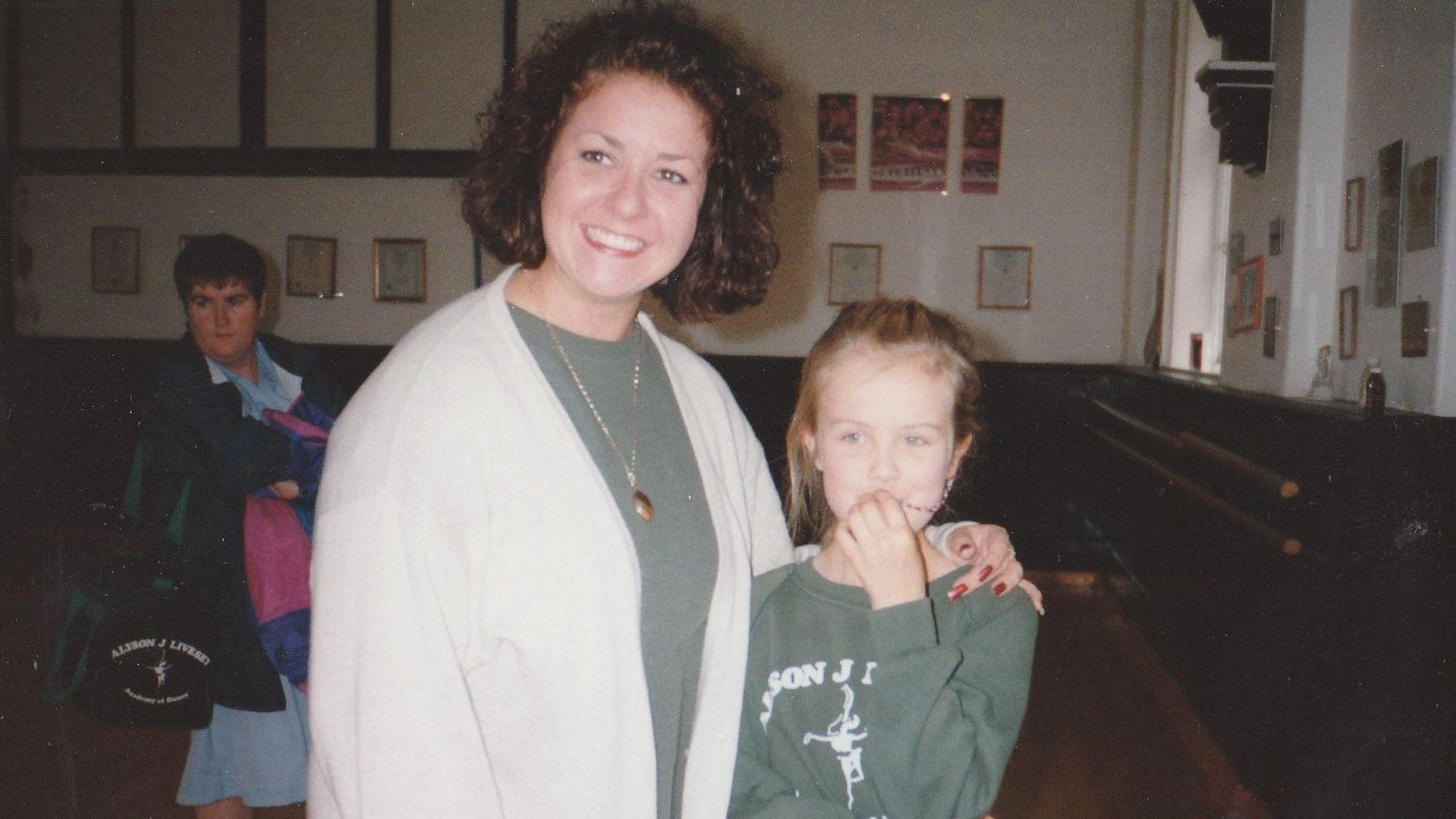 Claire Booth is pictured as a child in a dance studio wearing a green jumper next to Alyson Livesey who is has shoulder length hair and is wearing a cream cardigan and green top and a gold necklace.