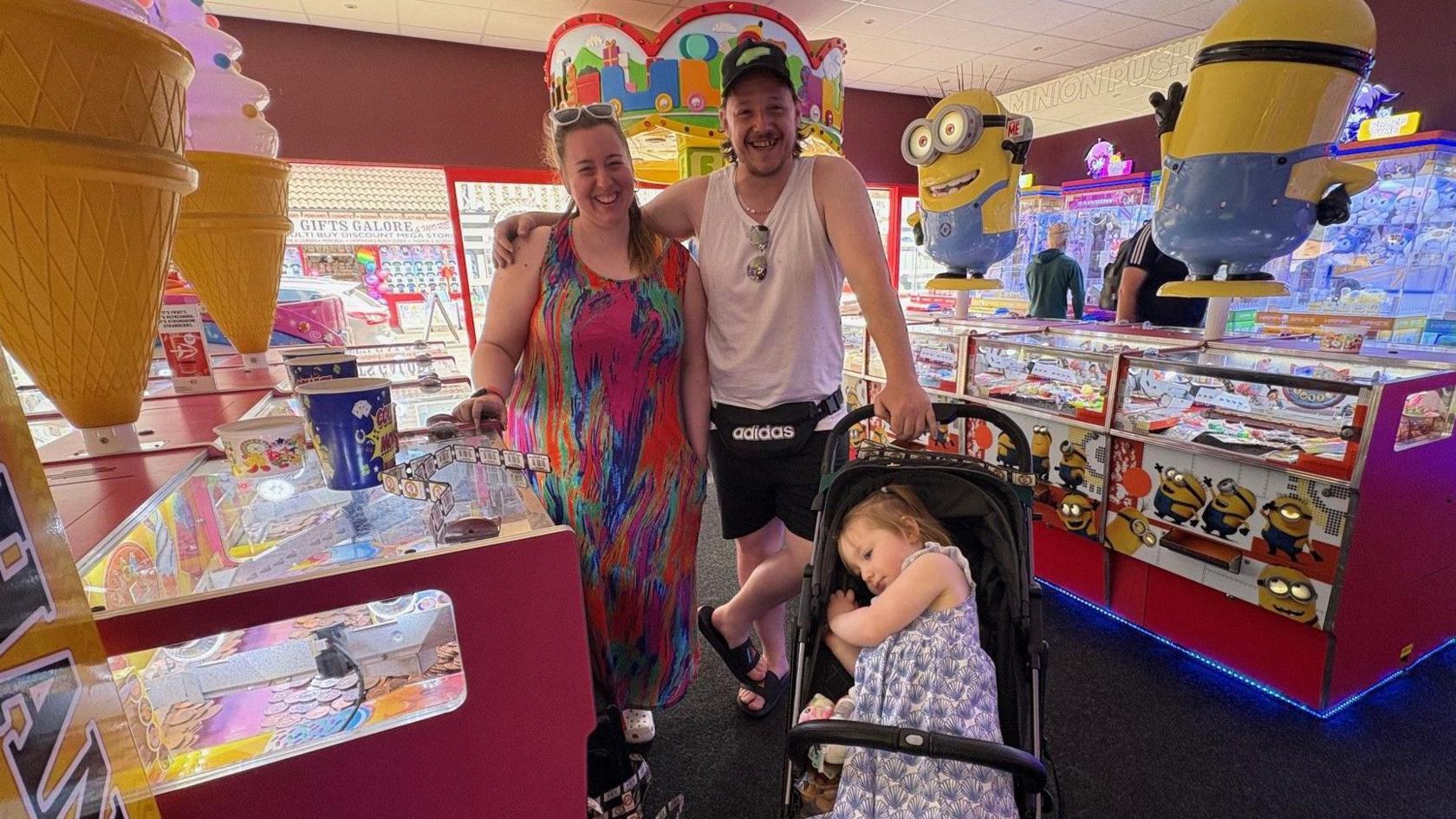 Stefan Careless, sporting a mullet hair style and wearing a vest top and shorts, is pictured with his partner Lucy Giacomin, who is wearing a multi-coloured long dress. Next to them is a pram containing their young daughter.