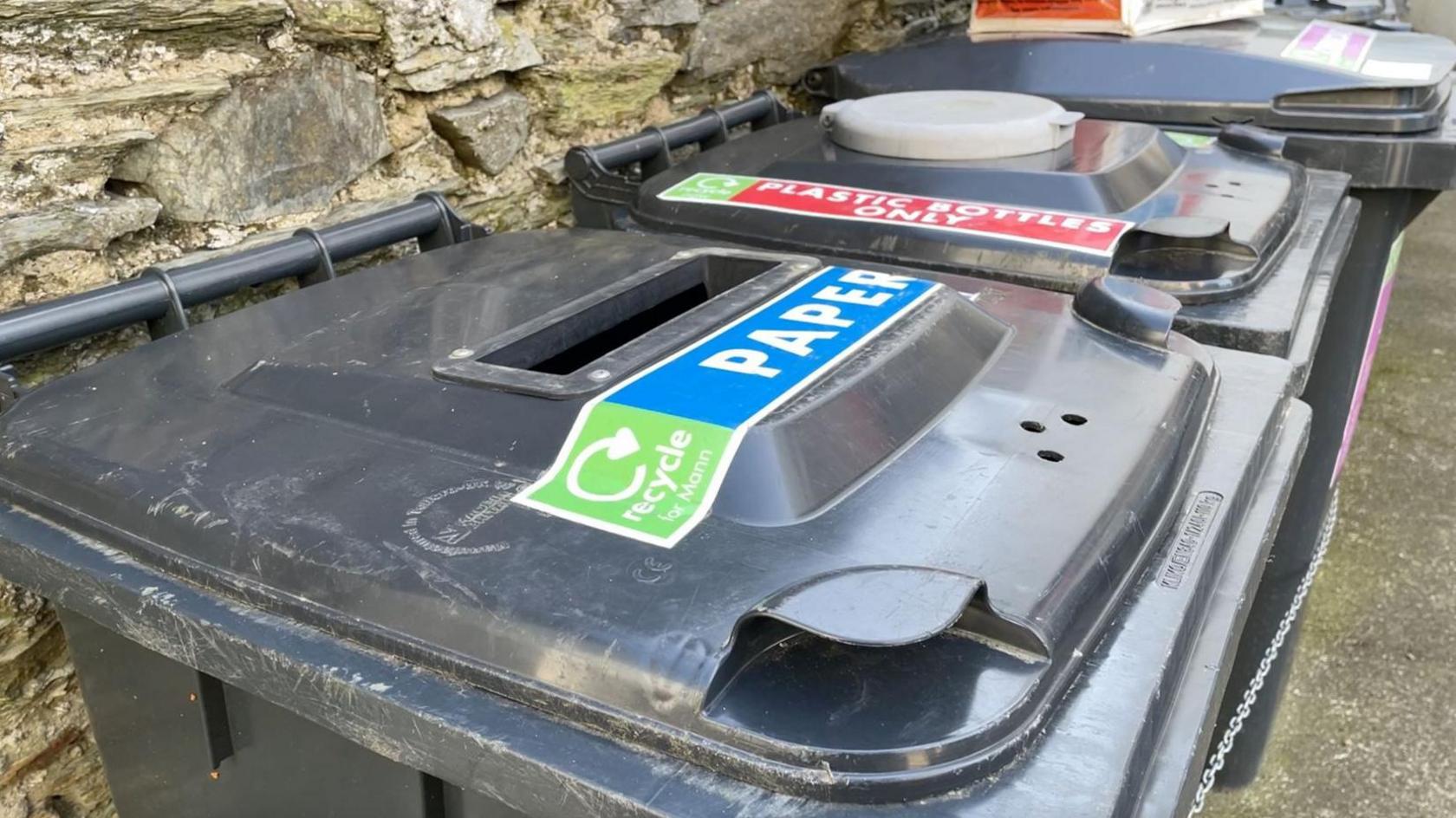 Paper recycling black bin in row of others against a wall.