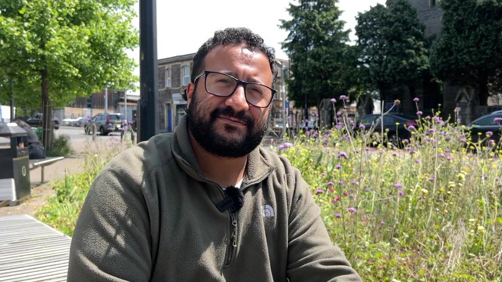 Courtney Young sitting on a bench nearby a road with a patch of grass and some flowers. He is wearing a khaki green fleece and has black rimmed glasses and curly hair.