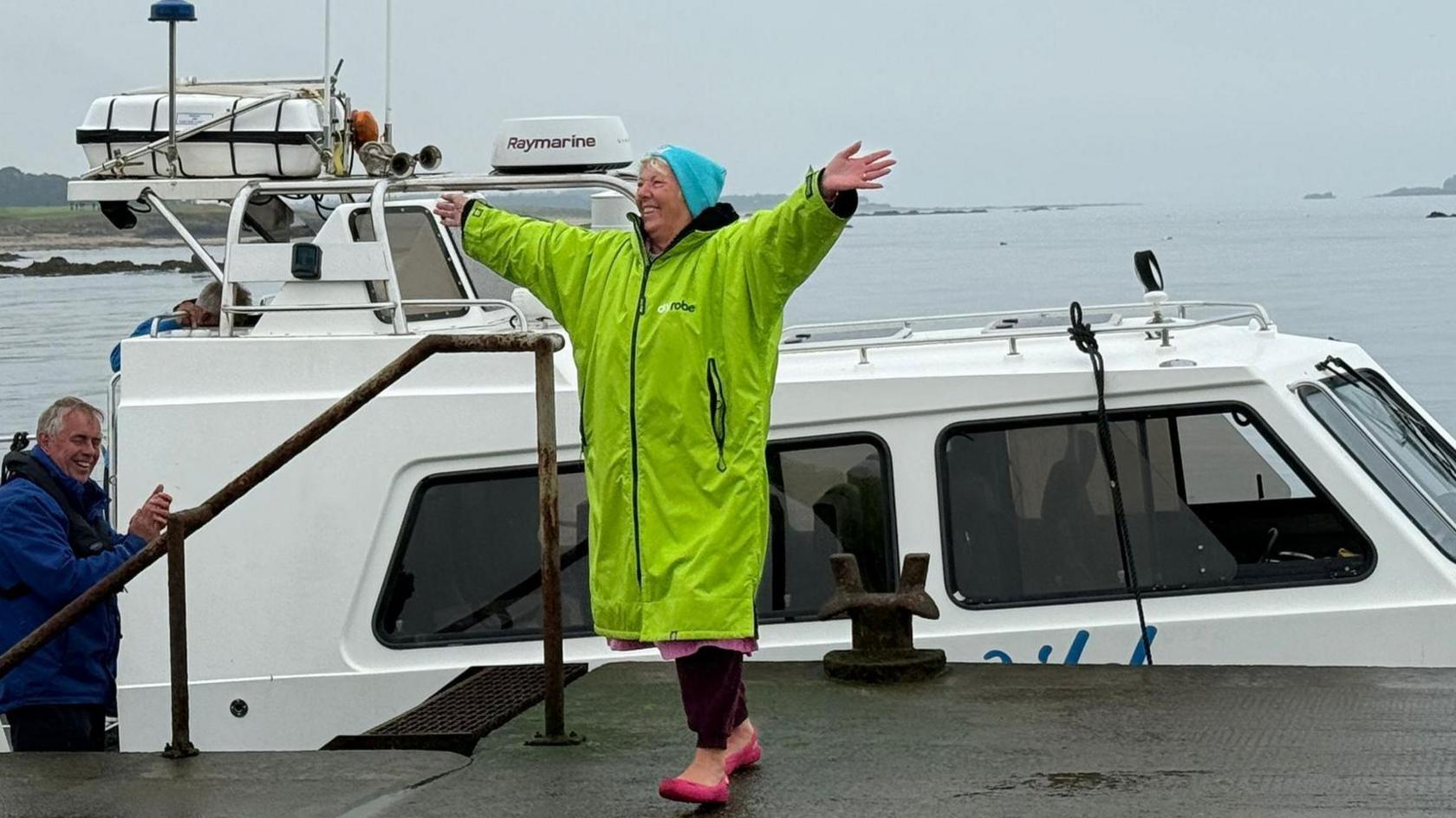 Woman in yellow dry robe and turquoise woolly hat, with her arms out-stretched. She appears to have just climbed off a boat, which can be seen behind her. 