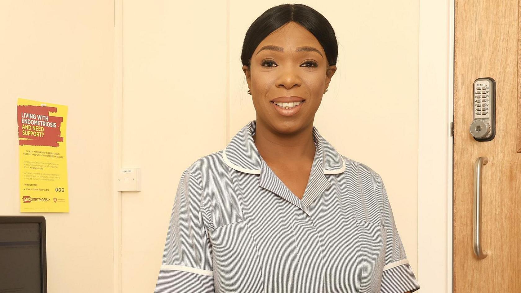 A woman is smiling at the camera, wearing a grey nurse's uniform