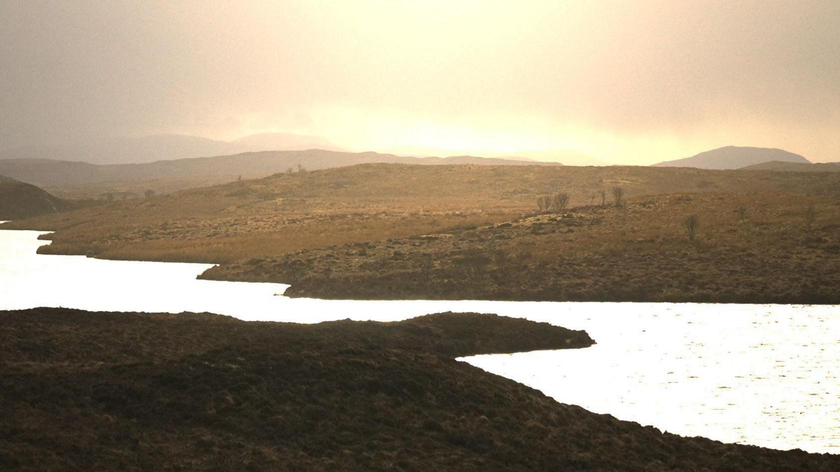 A rolling area of brown moor and peat bog with a loch in the middle of the scene.