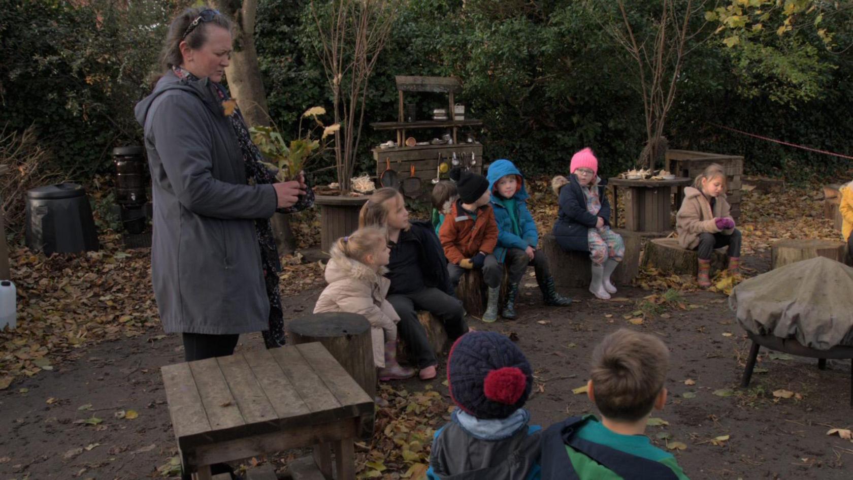 A group of eight pupils wearing coats and hats are sitting on upturned logs in a rural setting accompanied by a teacher holding the young oak tree who is standing next to a wooden table  