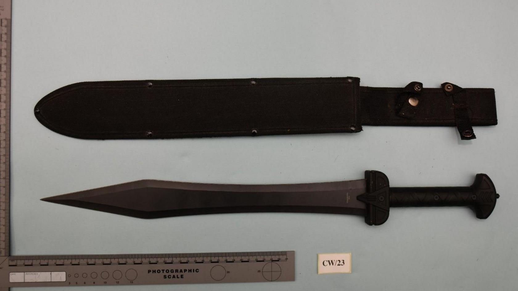 A sword placed on a light-coloured sheet next to a case used to store the weapon. There is a ruler next to the sword.