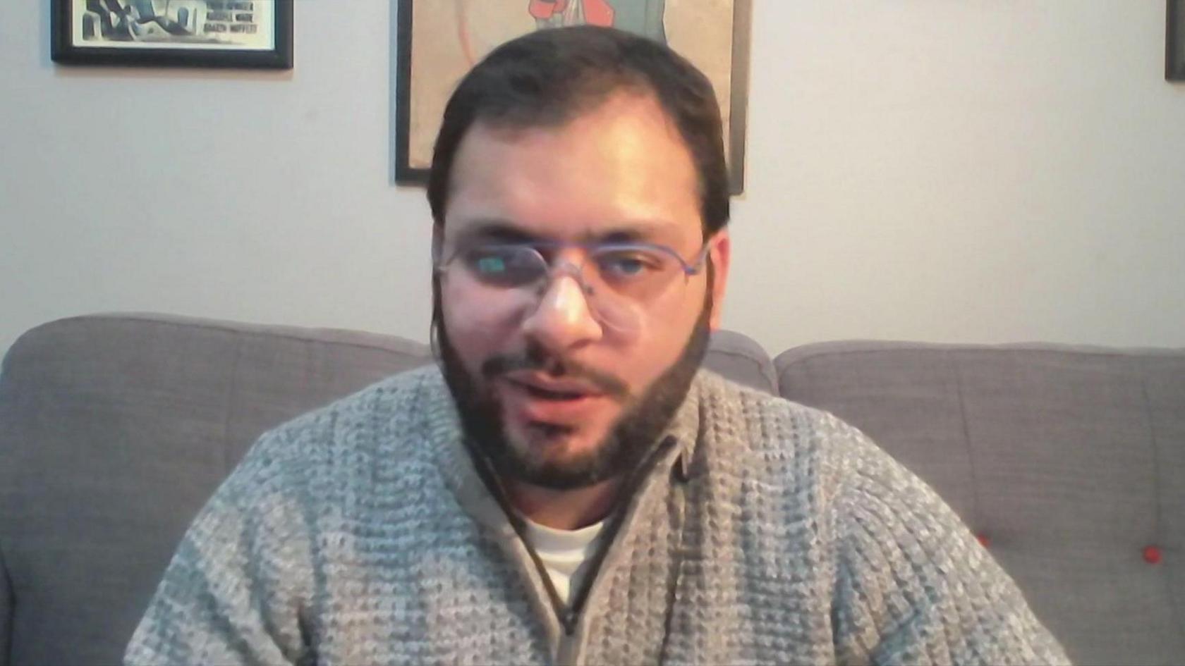 Avi Kherg looks at the camera whilst sitting on a grey sofa. He is wearing glasses and there is artwork behind him