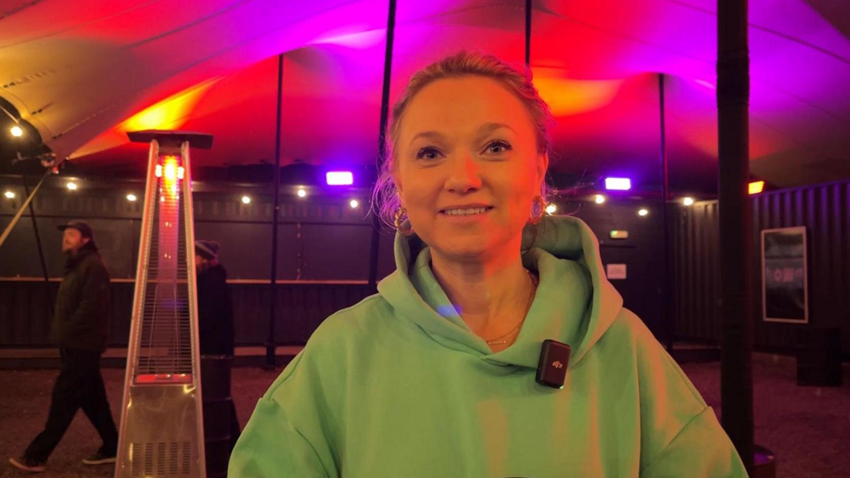 Bianca Mayhew is standing outside the venue under a lit up canopy. She is weating a green jumper and has her hair tied back.