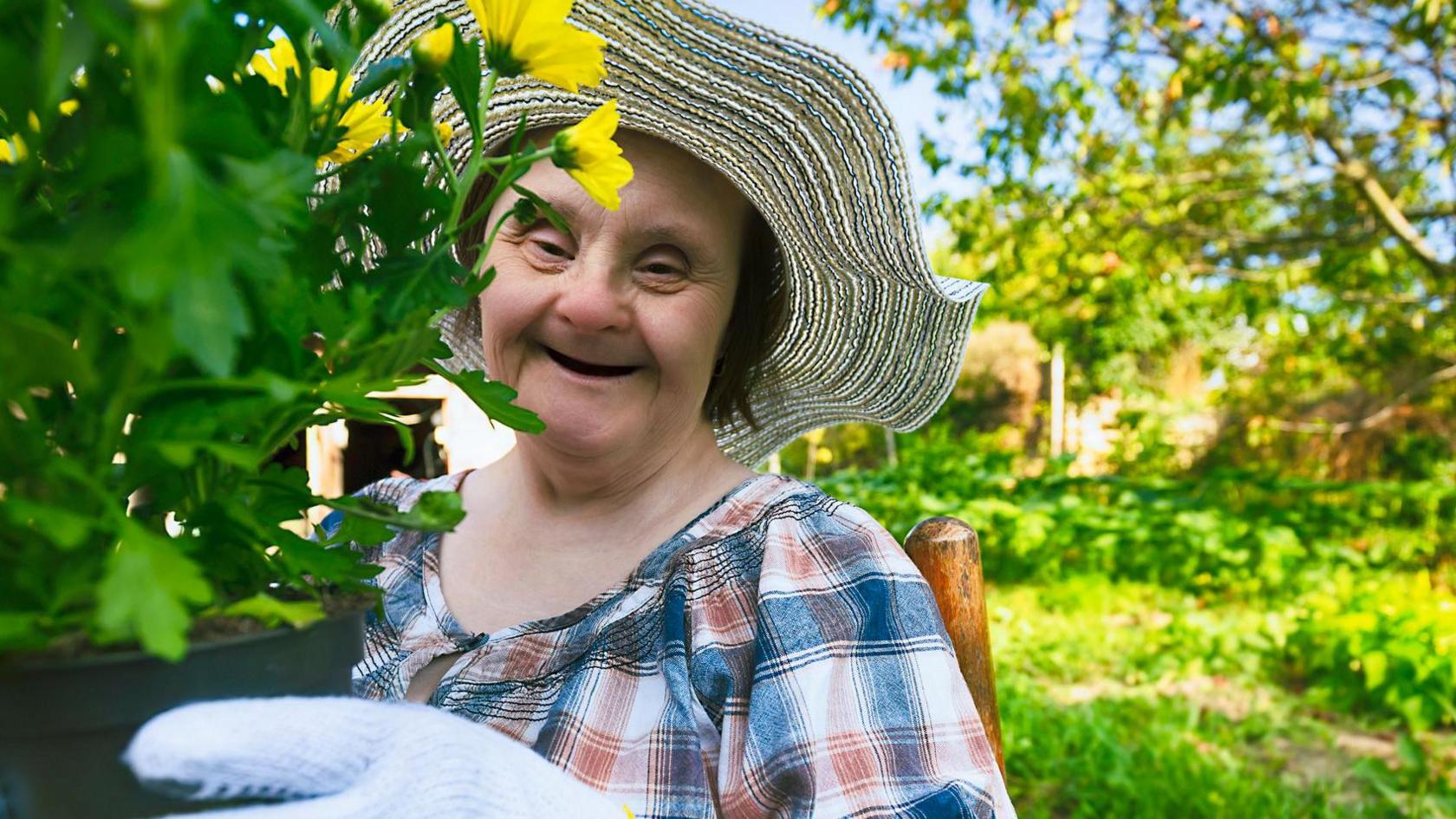 An older woman with Down's syndrome smiles at the camera while wearing a summer hat and holding a yellow flower in a garden (stock image)