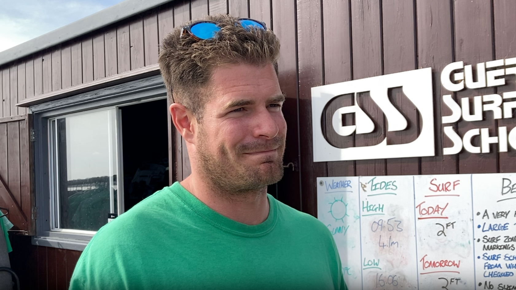 Nick Hill in a green T-shirt with brown hair. He is smiling and has sunglasses on his head. Behind him is a brown wood building with a half open slide door window, to the right a whiteboard depicting the tides, surf and weather. Above the whiteboard is a Guernsey Surf School logo in white