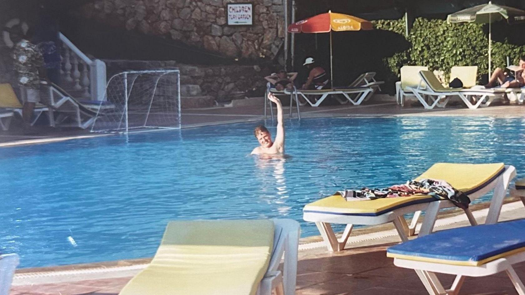 A young Jordan waves from a swimming pool on holiday. Sunbeds are in the foreground.