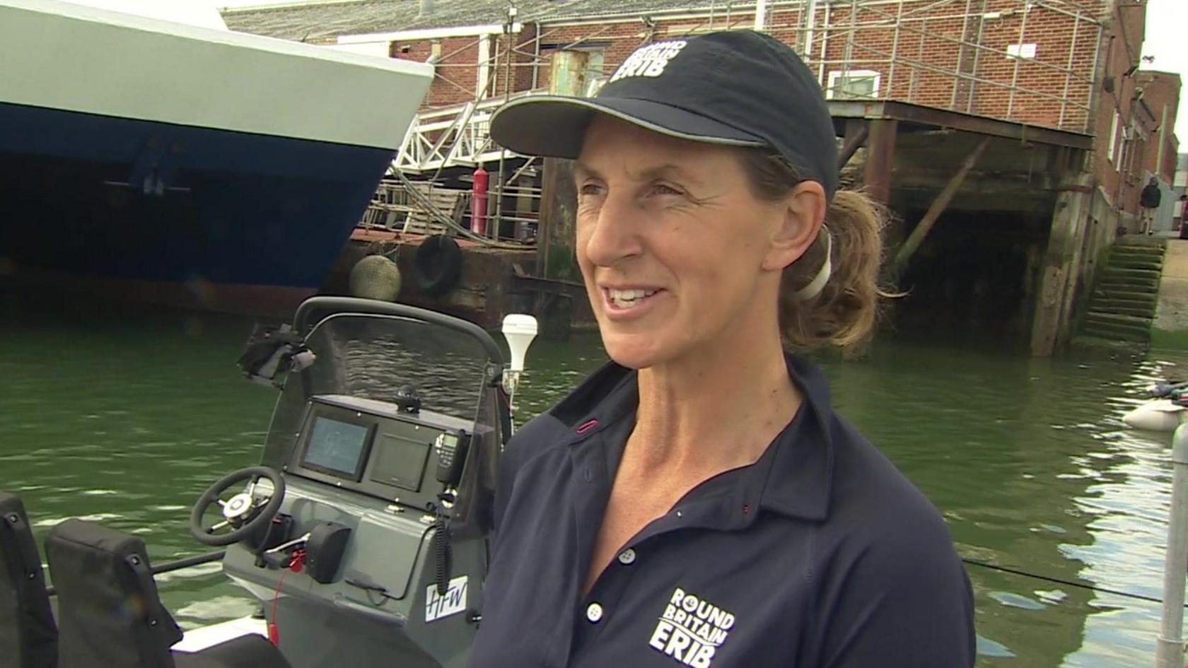 Jaqui Besley wearing a navy blue Round Britain ERIB t-shirt and cap stands in front of a boat in water