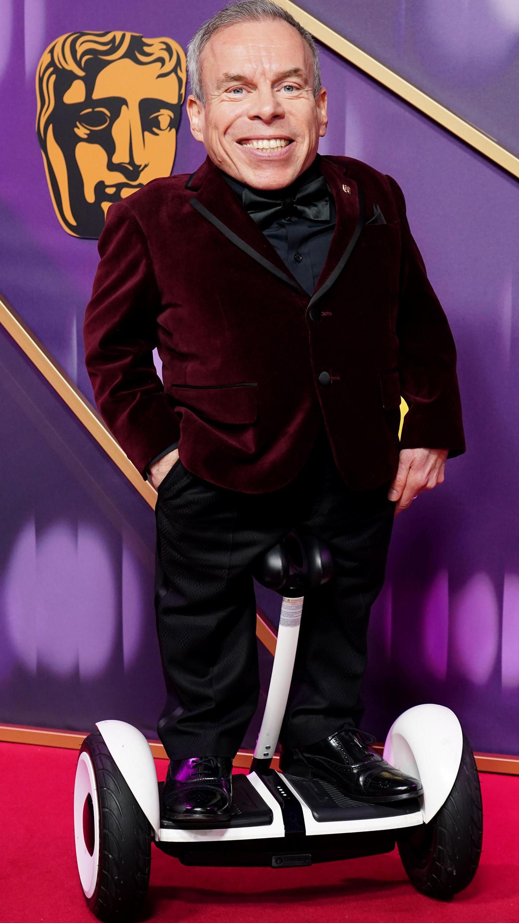 Warwick Davis in a maroon velvet suit on a motorised vehicle