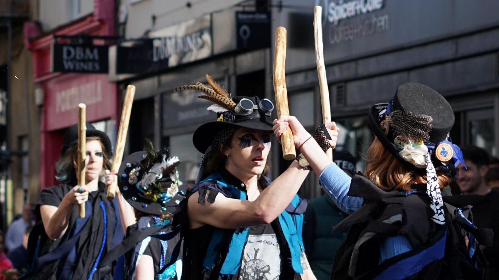 Sam Murphy dressed in black top hat with a feather, goggles, and a black and blue raggedy top, holding up a wooden stick with team members in view.