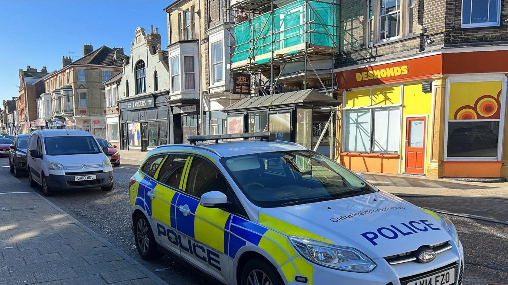 Police car parked on a street with shops and flats. Scaffolding can be seen on one building.