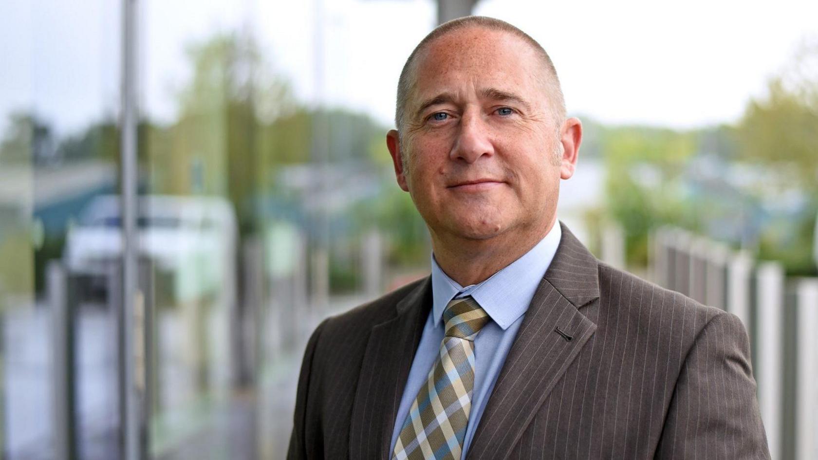 Detective Stuart Brooks, from Avon and Somerset Police, looks at the camera. He is wearing a grey pin-striped suit, light blue shirt and a cross-patterned tie.