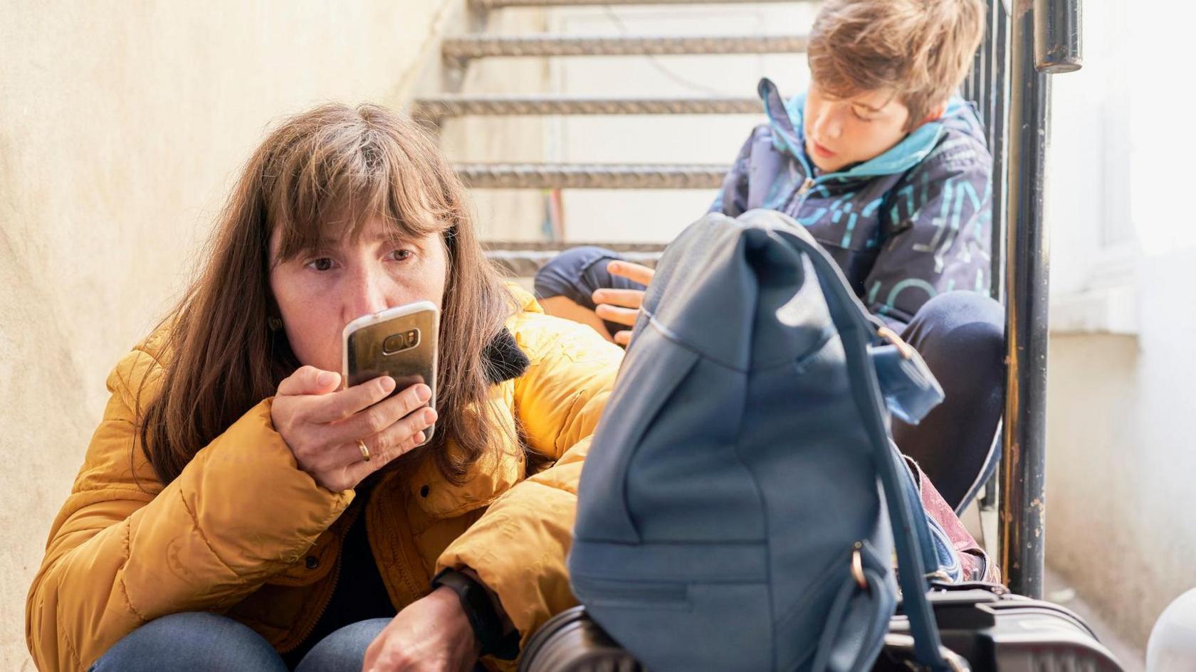 Mother sitting on a staircase, talking on the phone with suitcases and her children next to her