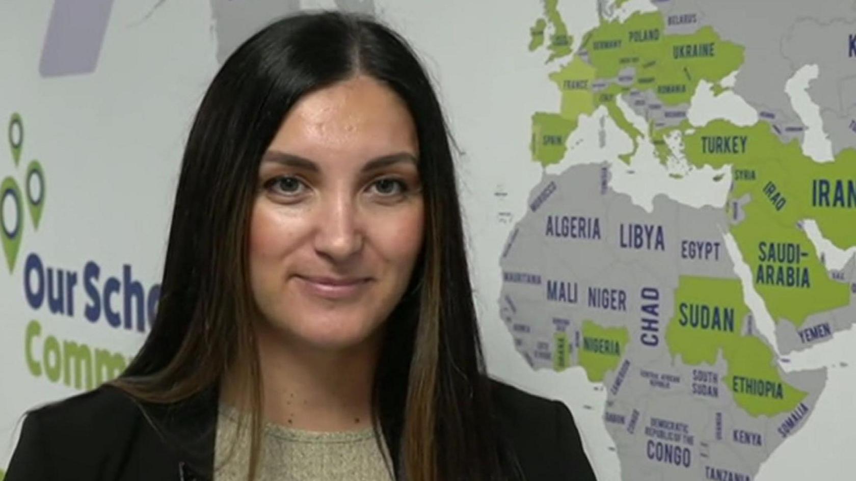 A woman with long dark hair wears a dark jacket with a lighter coloured top underneath. Behind her on the wall is a map showing many countries of the world