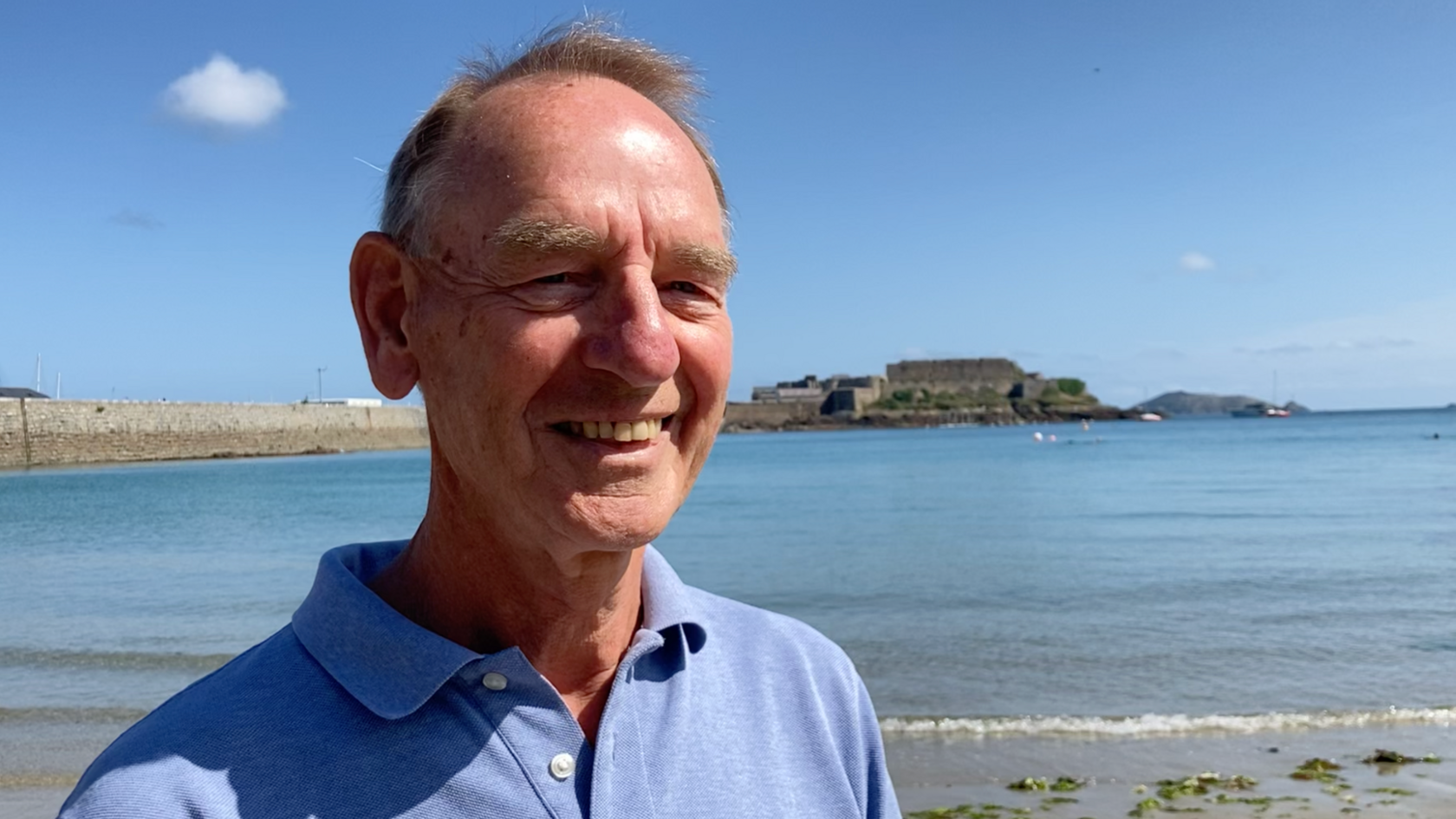 Paul Bugden standing on a beach. He has light brown hair and is wearing a light blue polo shirt. Behind him is the ocean with a pier in the distance