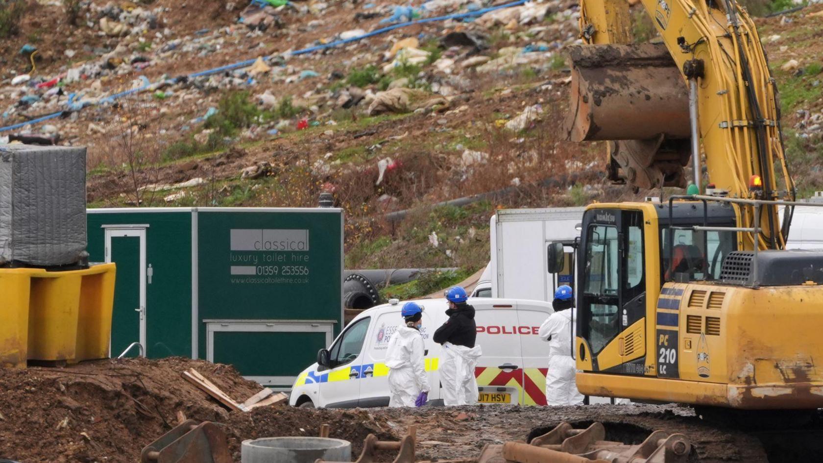 Forensic officers standing next to a digger at the landfill site in Stanway. Rubbish is strewn up a muddy hill where a search is taking place out of vision. There is a green portable toilet building and a white police van nearby.