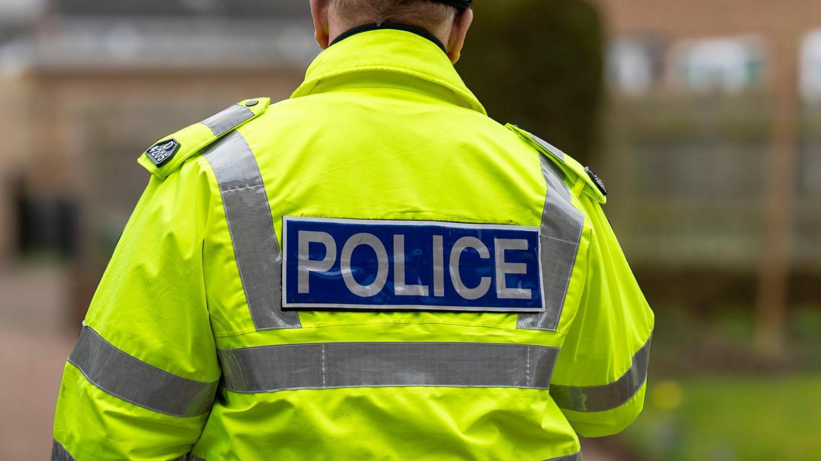 A policeman wearing a high-vis jacket, pictured from behind. Only his back and shoulders are visible. The jacket is in focus, while the background is heavily blurred. 