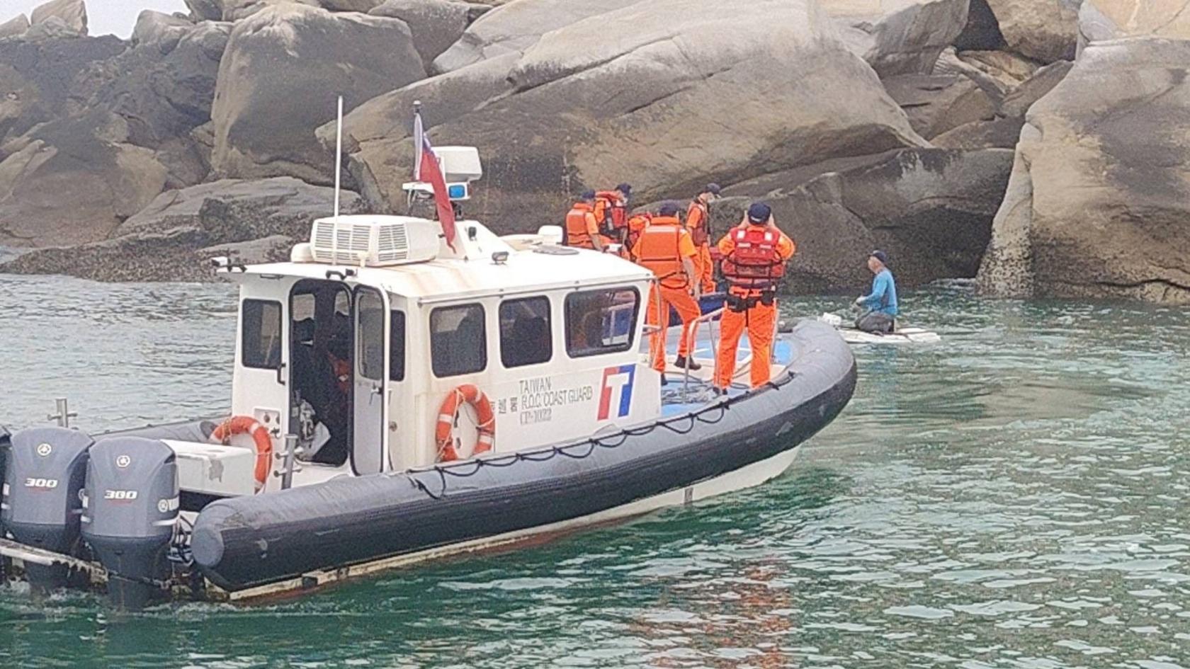 Taiwanese coast guard personnel dressed in orange uniforms on board a coast guard ship apprehend a suspect dressed in a blue top and grey trousers, who appears to be kneeling on a small floating platform. The vessels are by large rocks.