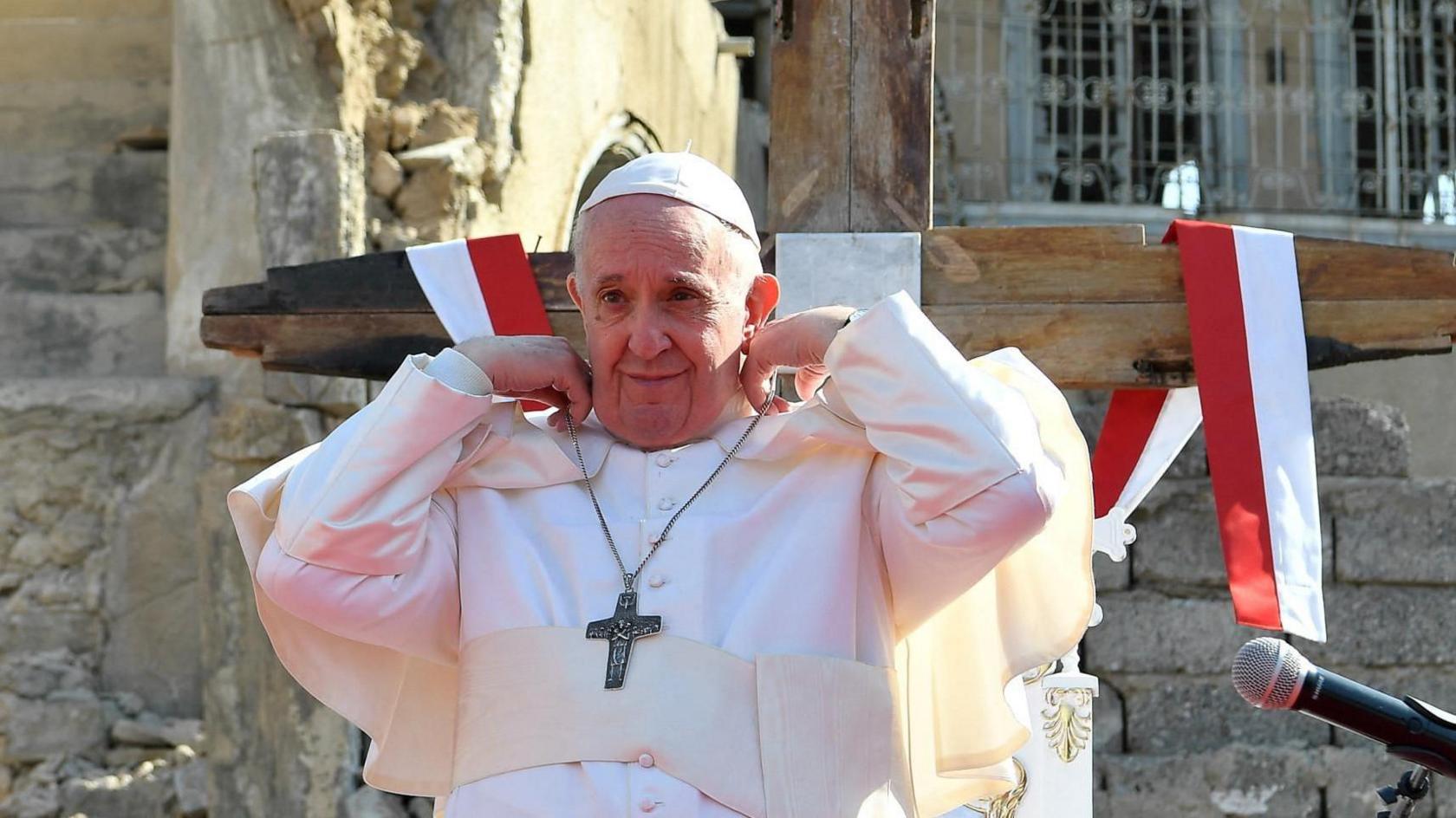 Pope Francis attending a prayer service in Mosul's Old City during a visit to Iraq in March 2021. He is seen adjusting a necklace holding a large cross around his neck. Behind him a large wooden cross can be seen. 