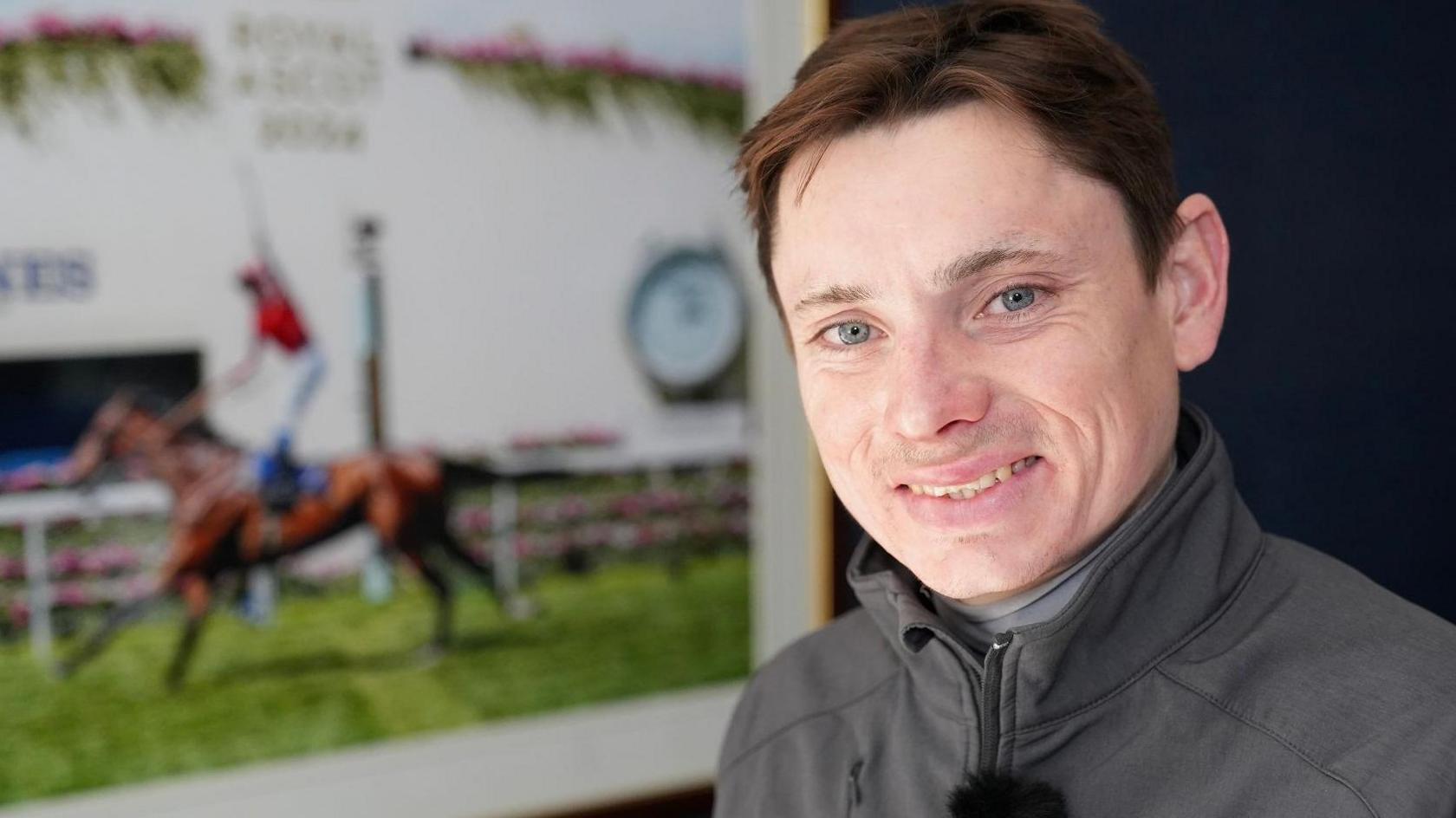 Callum Shepherd standing next to a mounted photo of his win at Royal Ascot last year