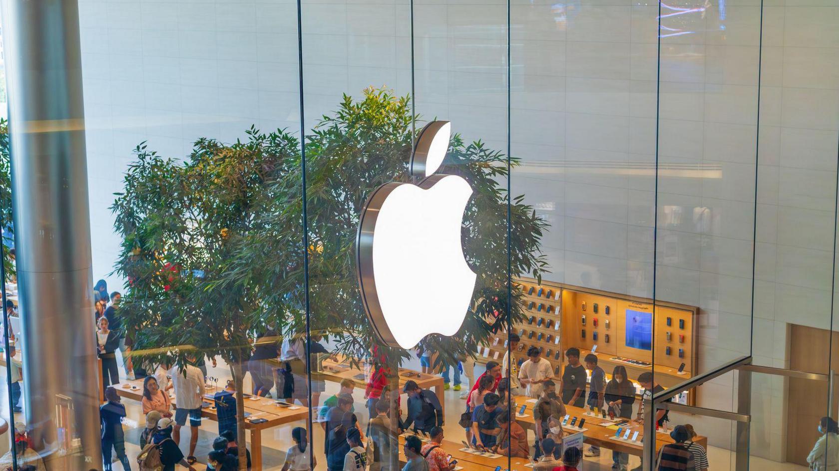 The Apple logo is pictured on the glass wall of an Apple store with people shopping below