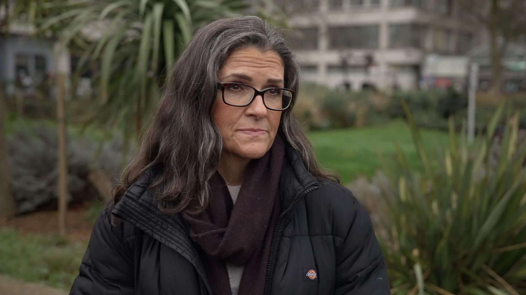 Jacqueline Wilson looks off camera in an outside shot with a hospital building and gardens visible behind her. She is wearing a black coat, dark-rimmed glasses and a dark coloured scarf. She has long dark hair.