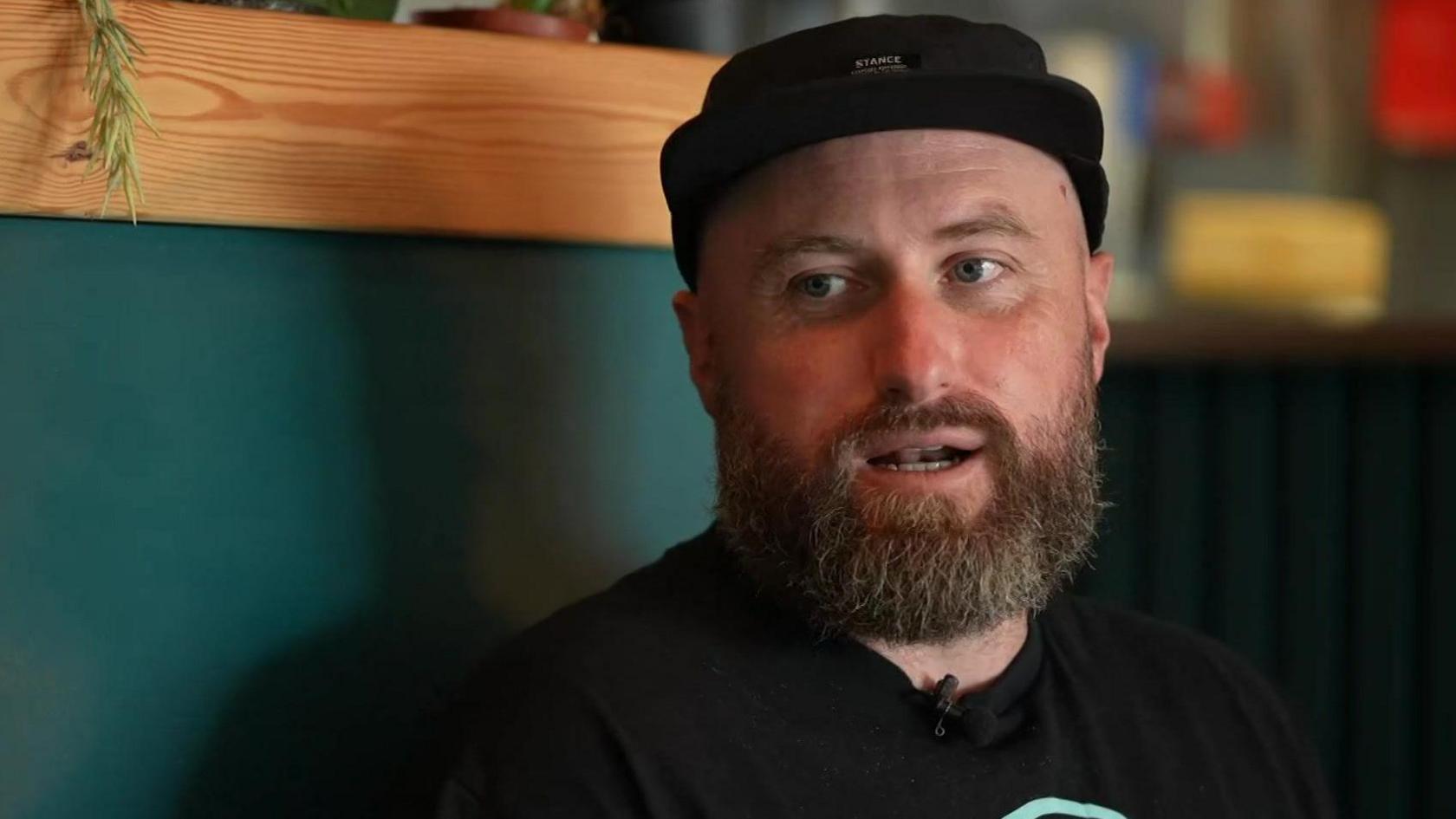 A bearded Oz Harborth wearing a black hat and black T-shirt. He appears to be talking to someone who is sitting beside him [out of camera shot]. Behind him is a blue wall.