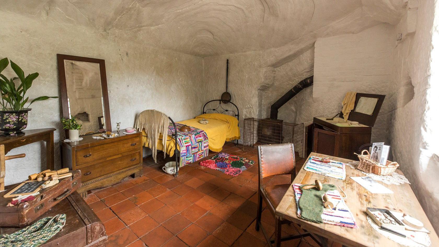 A whitewashed inside of a home with reddy tiles on the floor. An iron bed stands in one corner with a dark wood chest of drawers next to it, with a mirror on top. Other furniture including a table and chair are in the room. a pipe from a fire stove can be seen next to the bed.