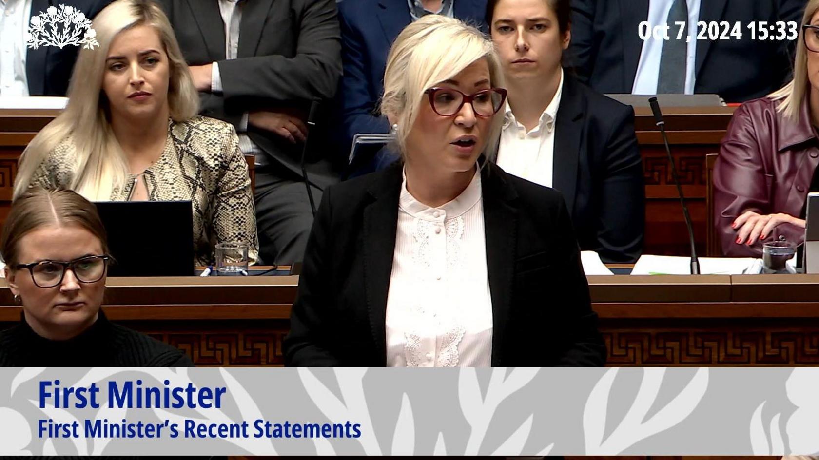 Michelle O'Neill - a woman with blonde hair in an updo wears red-framed glasses. She is standing with other MLAs around her and she is wearing a black suit jacket and white buttoned shirt.