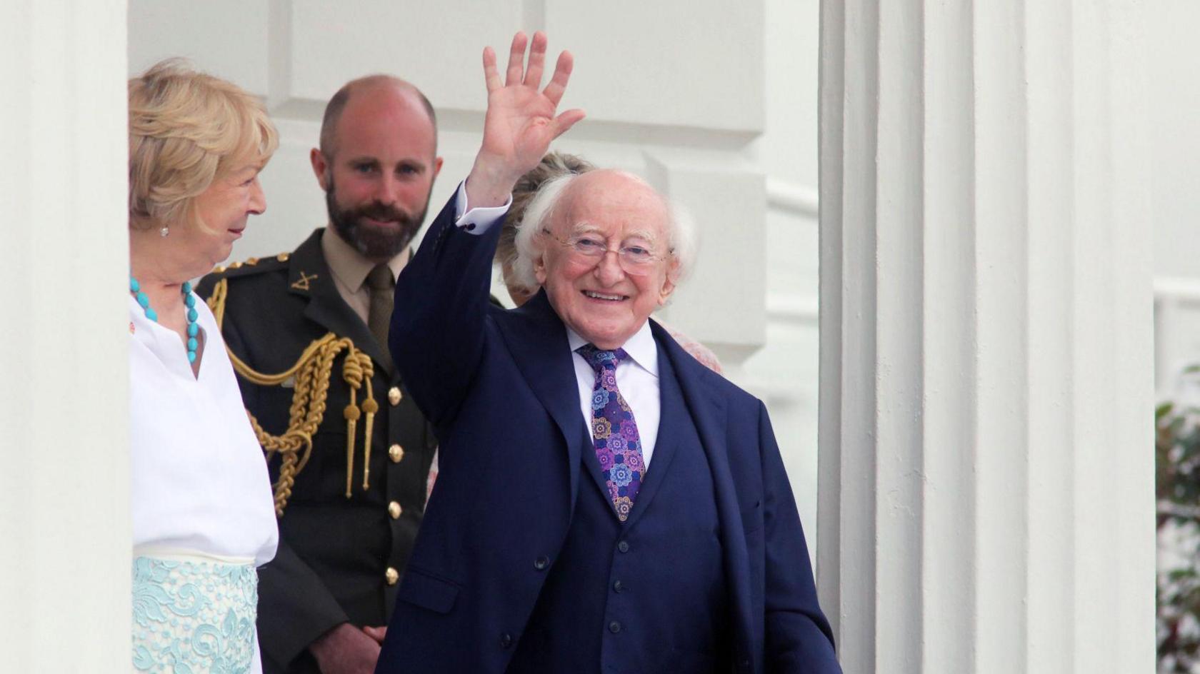 Michael D Higgins waves to waiting press. He is balding at the top with white hair down the sides. He has a navy three piece suit on and a flowery tie. Sabina stands beside him. She is in a white shirt, turquoise necklace, white shirt and white and turquoise skirt. They are on the steps of Aras an Uachtarain. Behind them stands a bald man, with a brown beard, in an official uniform.