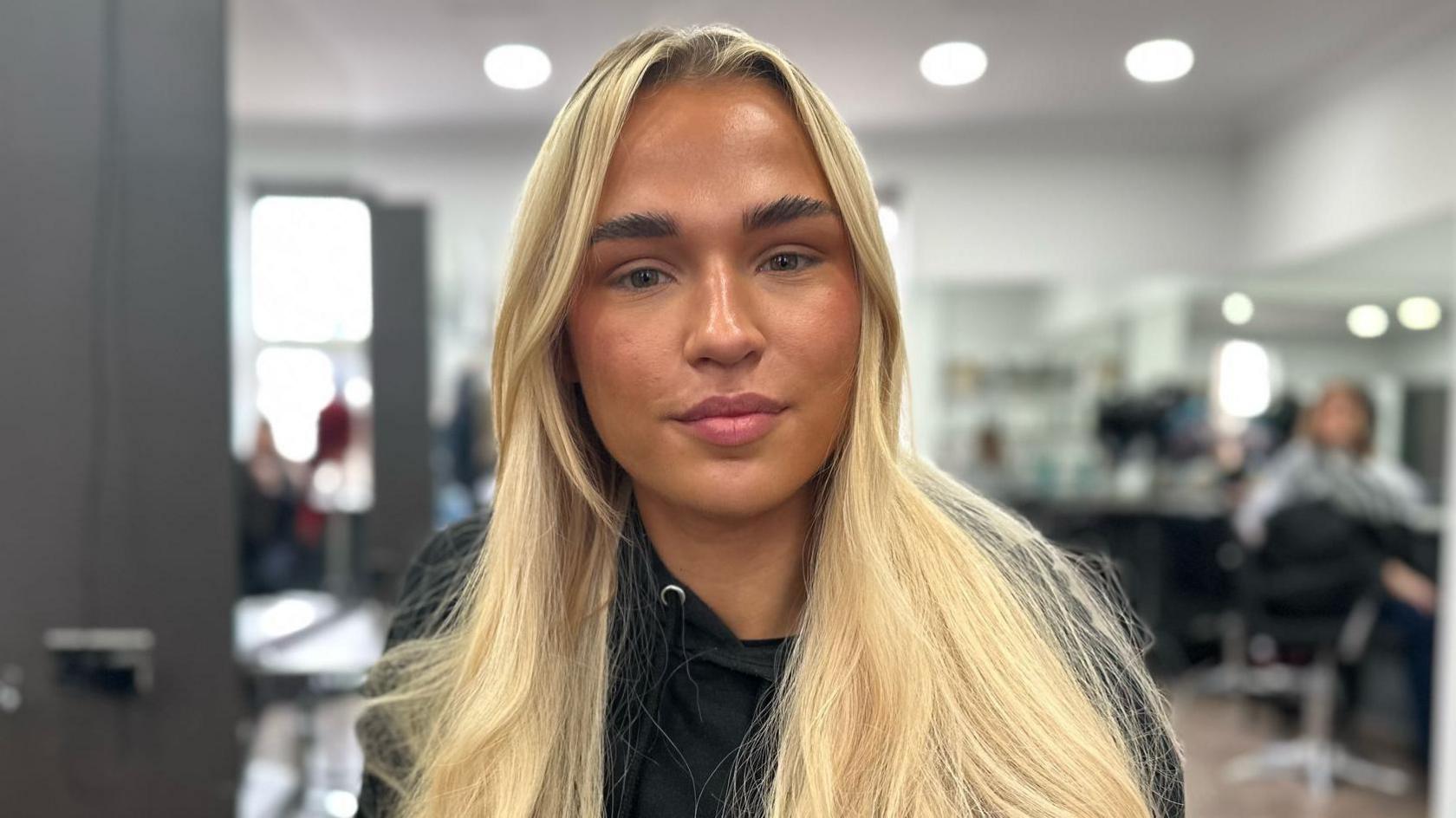 Alex Ritchie pictured in a salon. She has long blonde hair worn loose and wears a black hoodie. Behind her are salon chairs, mirrors, and clients getting their hair cut. 