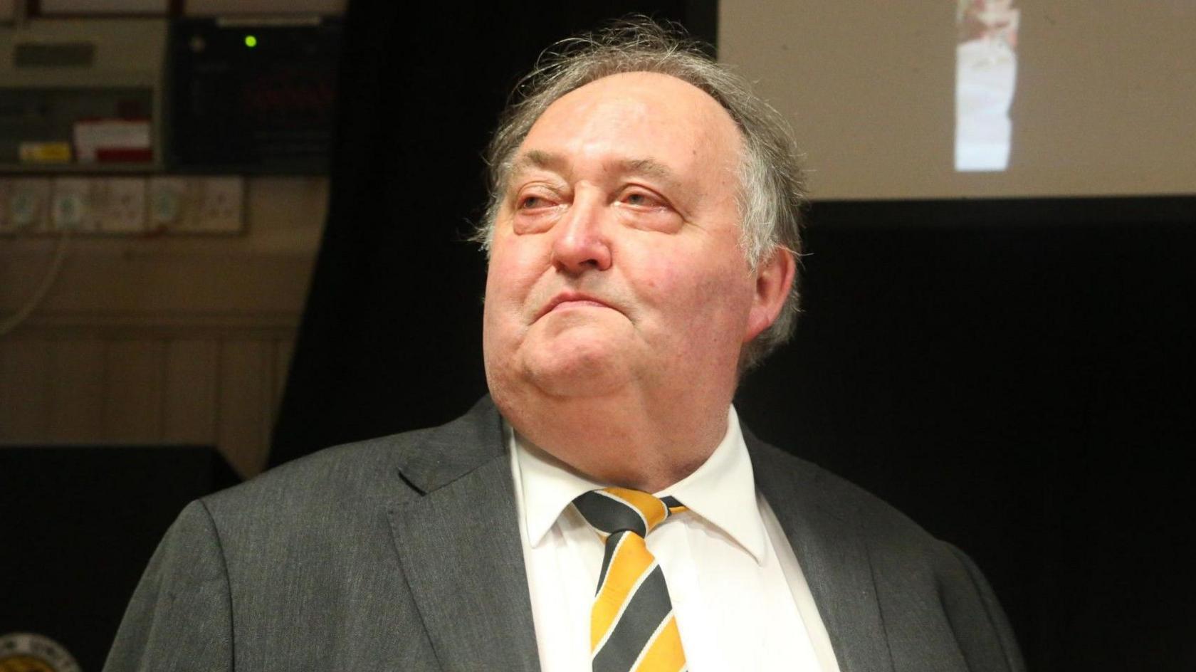 A head-and-shoulders shot of a man wearing a shirt, a yellow and black tie and a grey suit jacket. He has grey hair and is looking away from the camera.