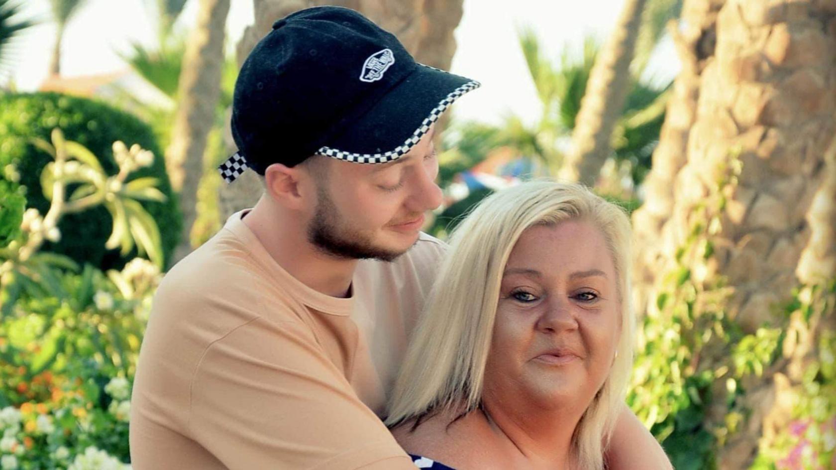 Joe Nihill on the right, wearing a black cap and beige t-shirt. He has his arms around his mum, Catherine, and is looking at her and smiling. She is also smiling, and looking at the camera. She has blonde hair. They seem to be on holiday, with the blurry background seeming to contain palm trees and a few umbrellas.