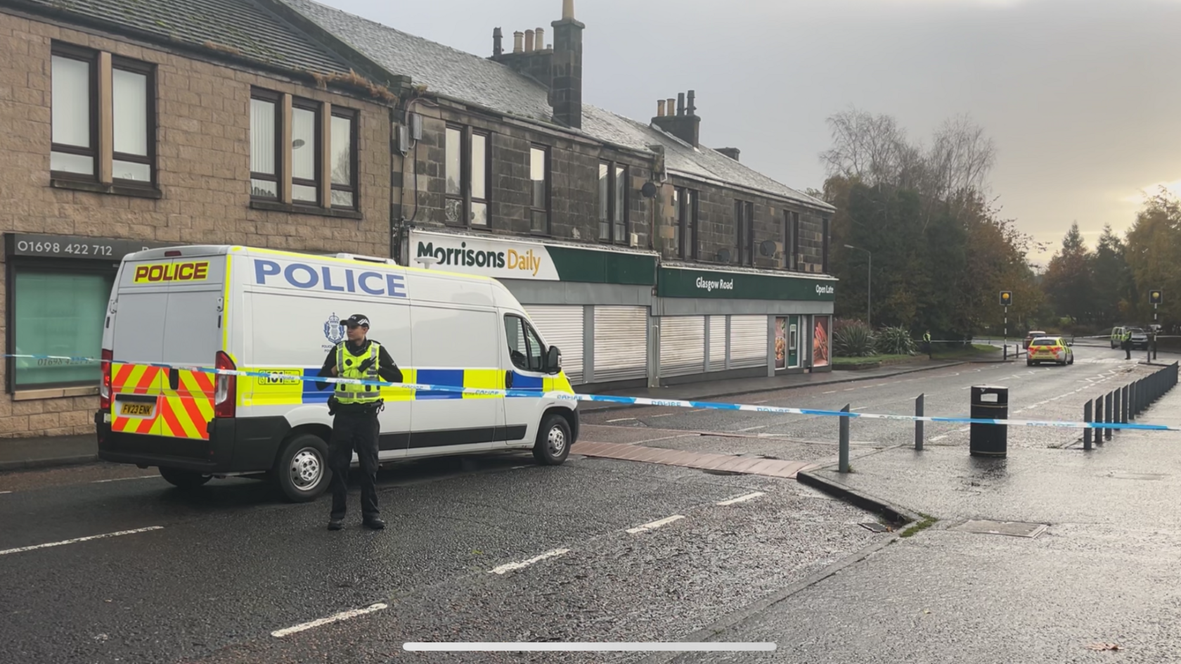 A street in Hamilton with a large police van parked on the road outside a branch of Morrisons. There is a police officer standing in front of the van looking out of shot to the left of frame. The weather is wet and cloudy