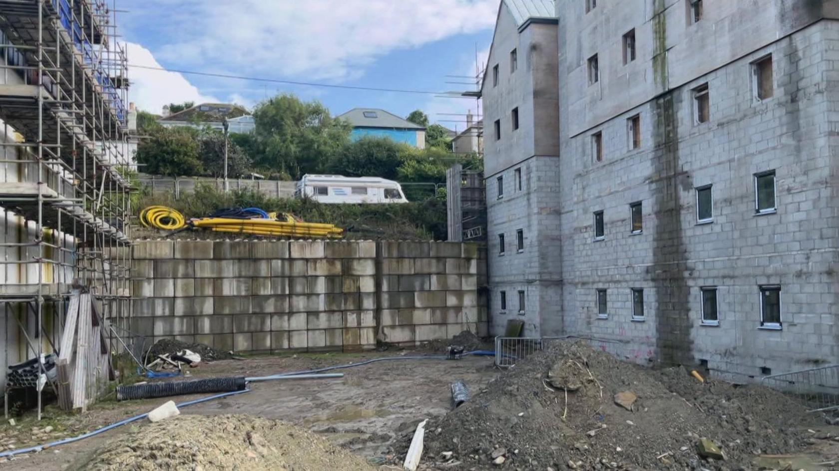 A building site. A wall of a block of flats is unfinished with the breeze blocks visible. On the ground there are piles of earth and building rubble.