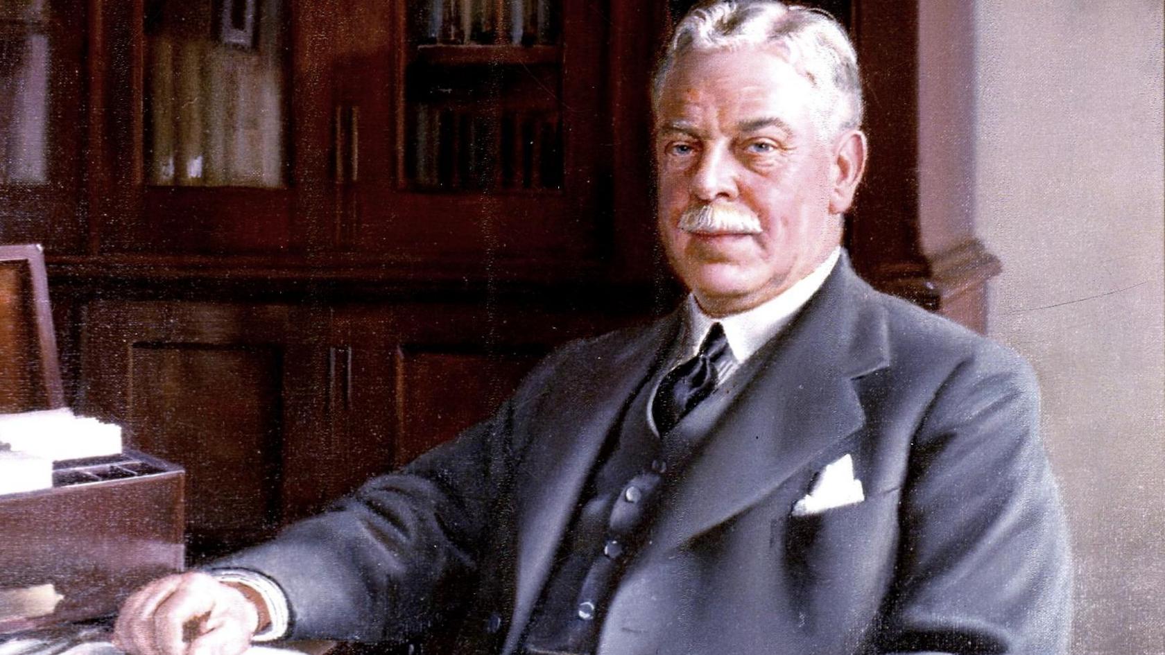 A colourised portrait of Sir Nigel Gresley, who is wearing a three-piece grey suit and sitting a desk in front of dark-wood bookcases. He has short grey hair and a moustache. 