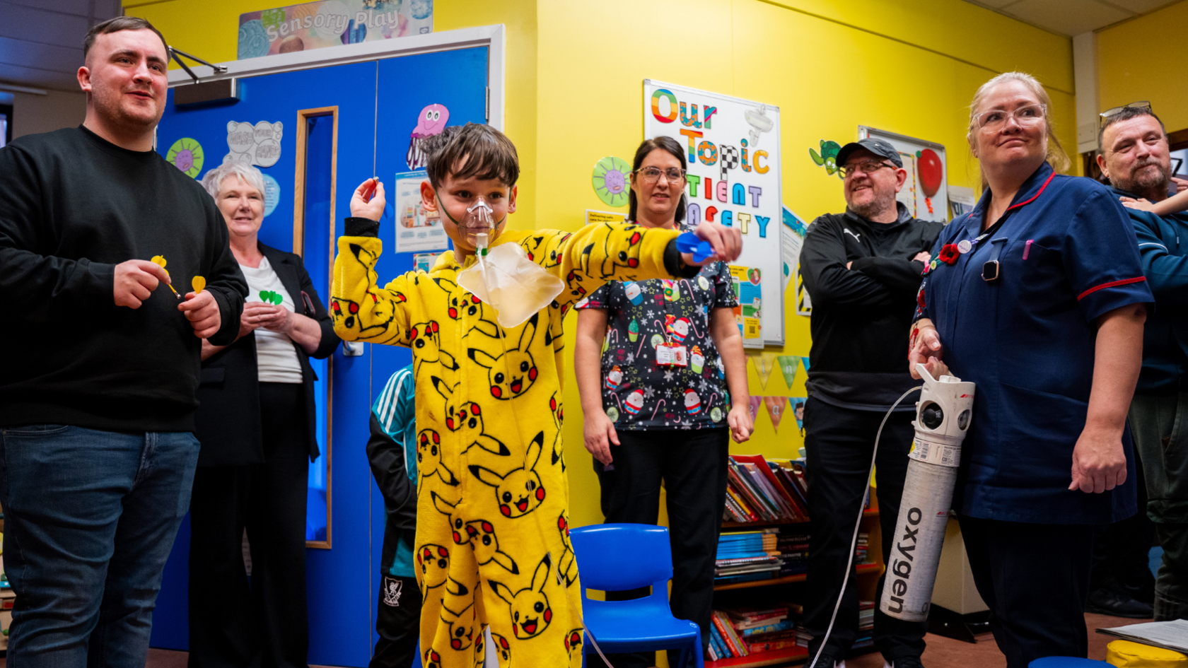 A child in a bright yellow Pikachu onesie stands in a hospital playroom, arms raised throwing a magnetic dart. Luke Littler is behind him holding several more magnetic darts. Several adults and staff members stand nearby, some holding small toys or medical equipment. The walls are decorated with posters and learning materials.