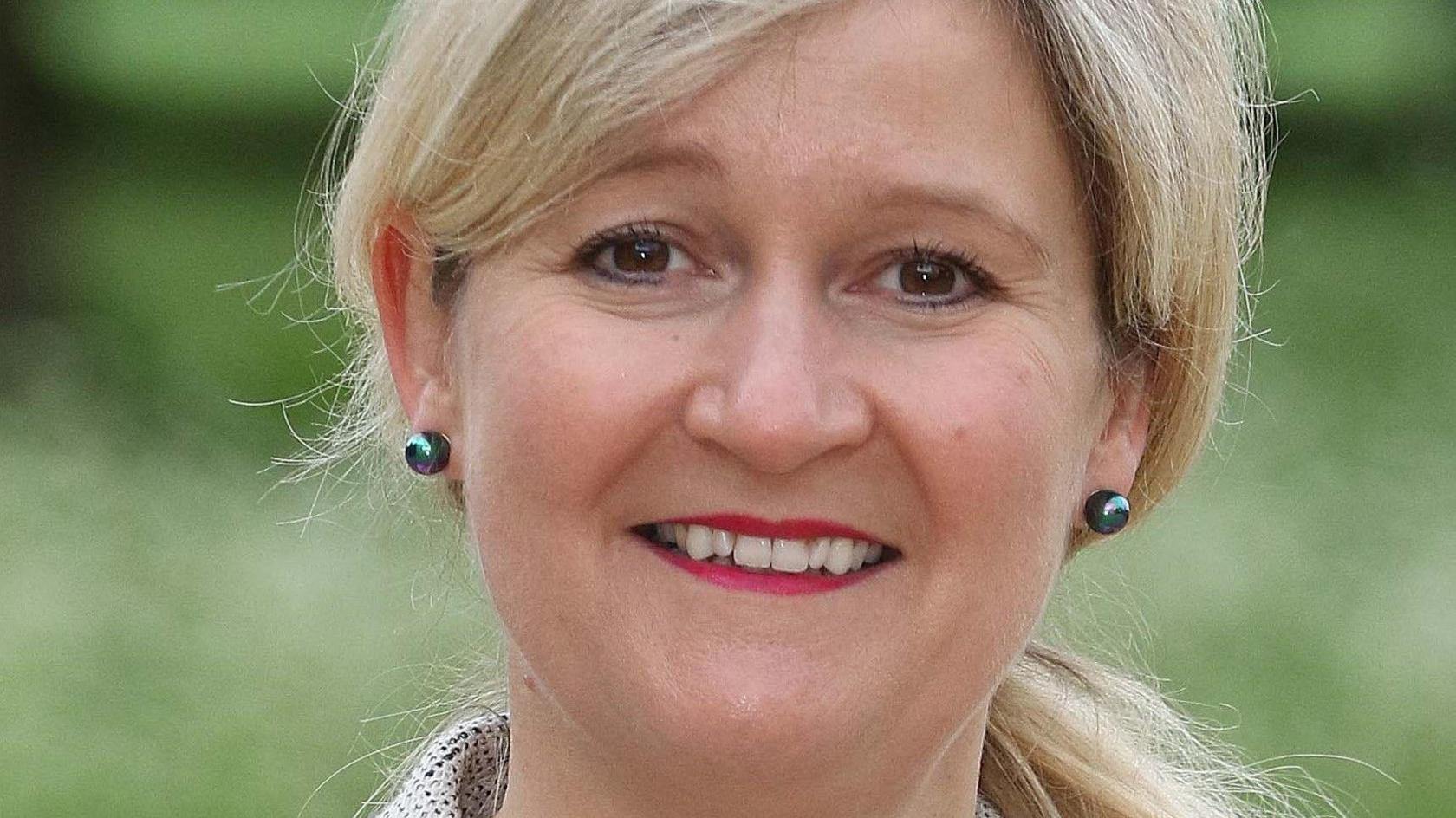Amanda Lyes looks at the camera and is wearing rounded metallic earrings and pink lipstick. She has blonde hair tiedback and the image shows her face close up