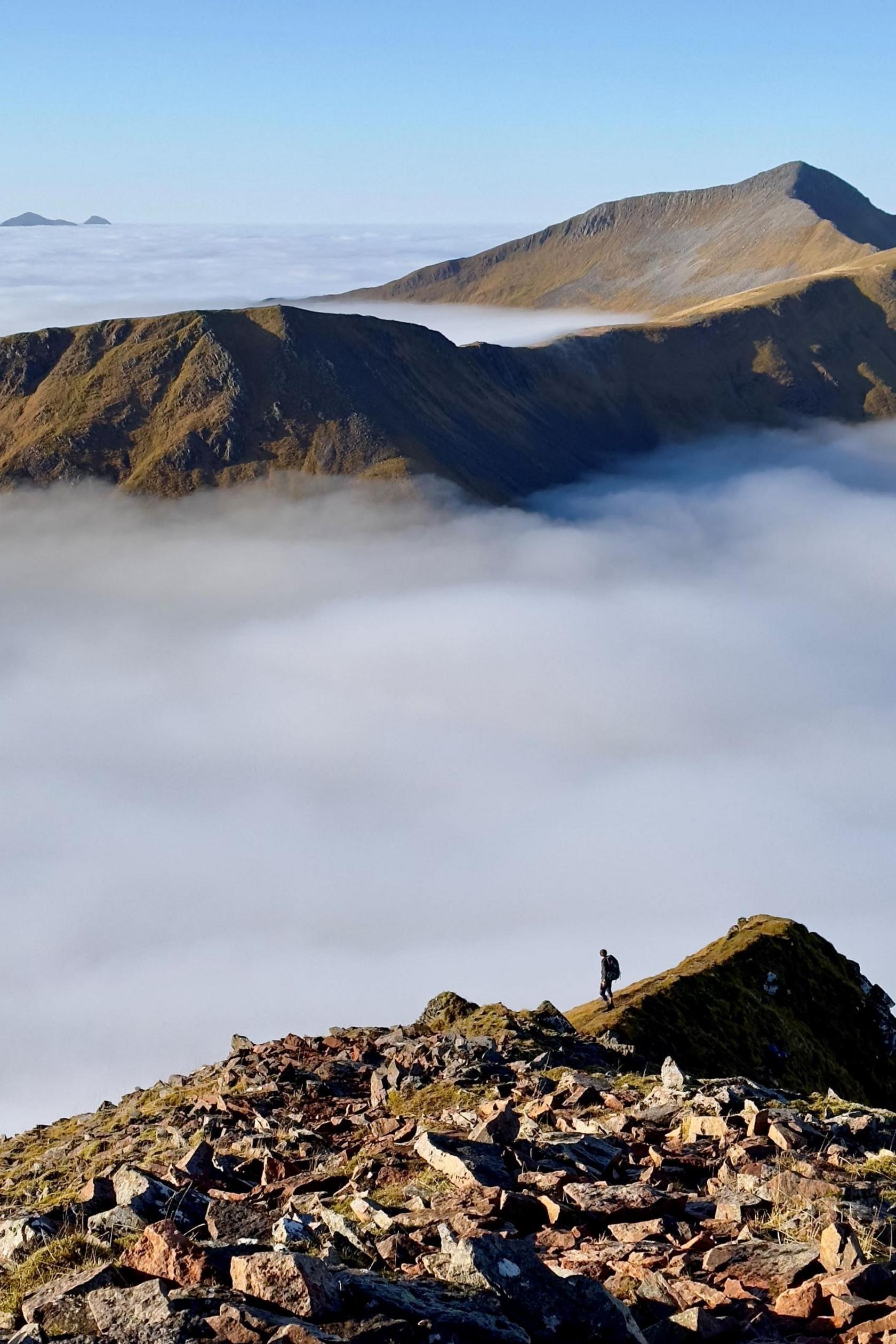 A hilwalker descends a slope on a mountain. They are silhouetted against white cloud. Other mountain tops appear above the clouds.