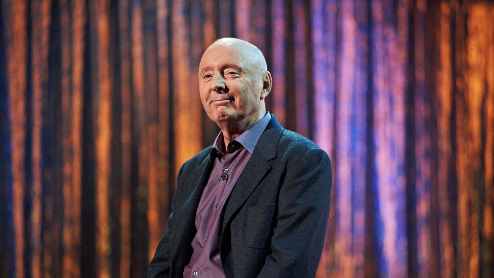 A man sits in front of a shiny stage curtain. He is bald, and he is wearing a dark suit jacket and a purple shirt.