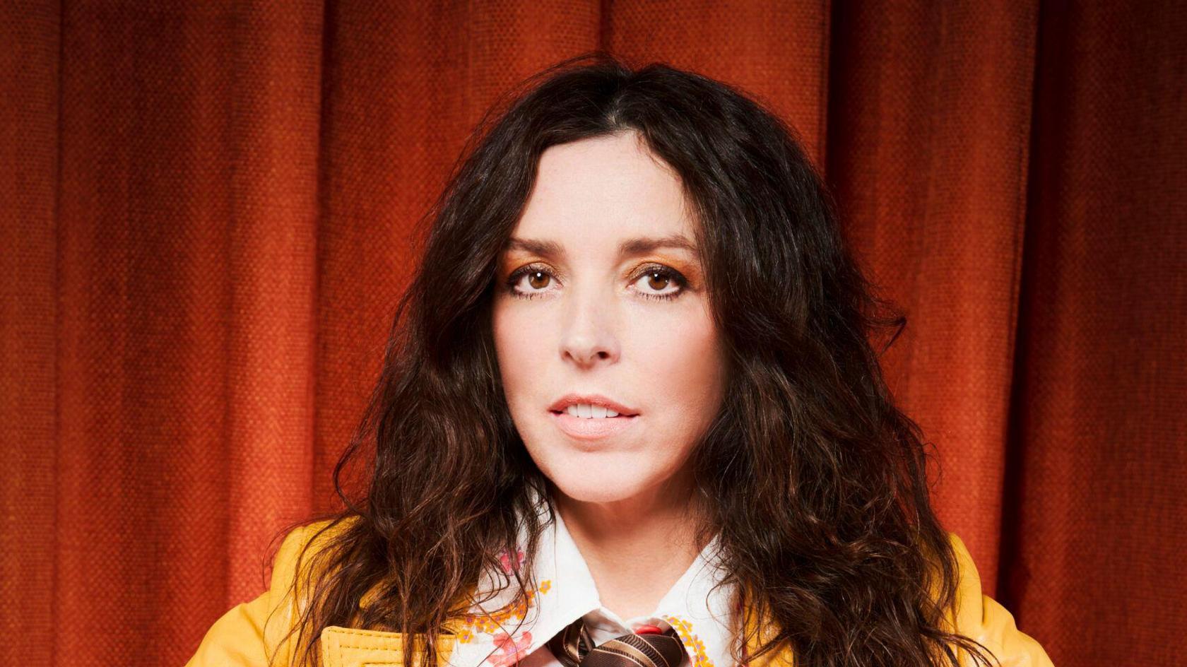 A woman smiles at the camera in front of a red curtain. She has shoulder length and wavy brown hair. She is wearing a yellow jacket with a white shirt underneath with a floral pattern on it as well as a tie.