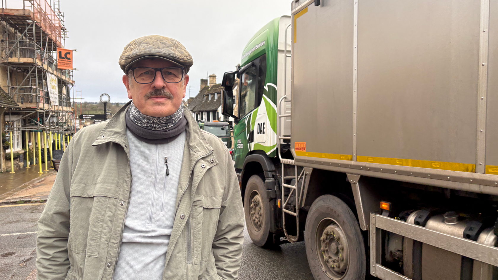 Paul Newman stands at a junction in the middle of Burford as a lorry passes. He has a moustache and is wearing a cap, glasses, a snood and a light-coloured jacket over a white top.