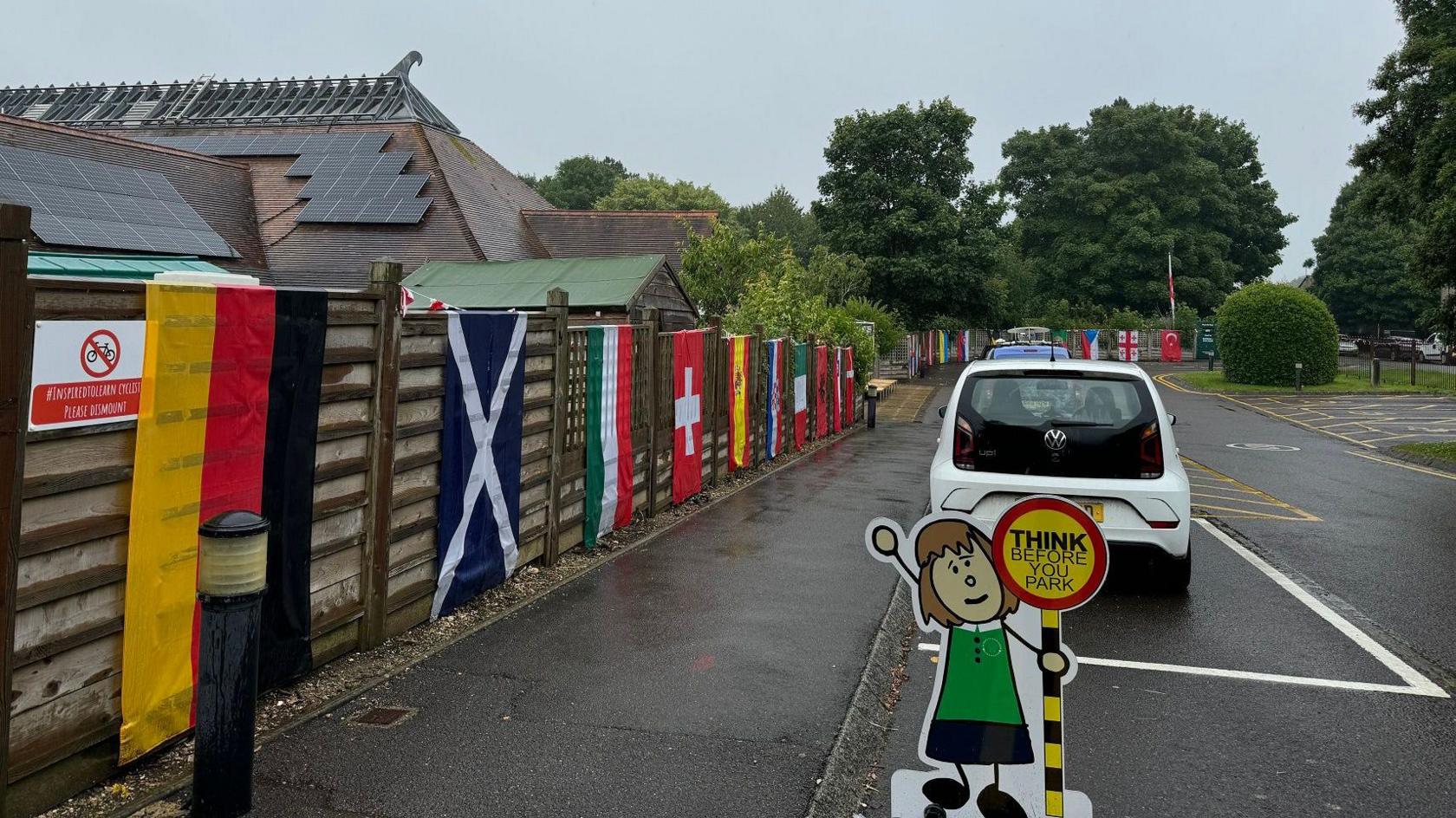 A school fence with a variety of European flags draped over it, next to a school car park