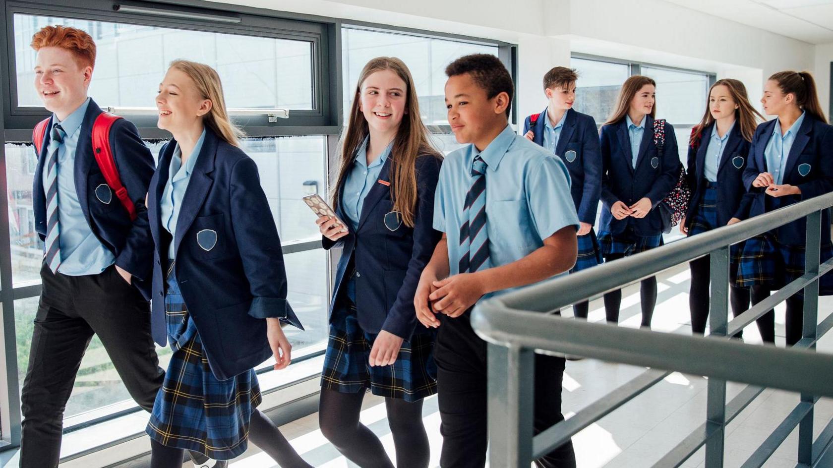 A group of High school students walking to their next class. There is a mix of eight boys and girls and they wear blue and tartan uniforms. 