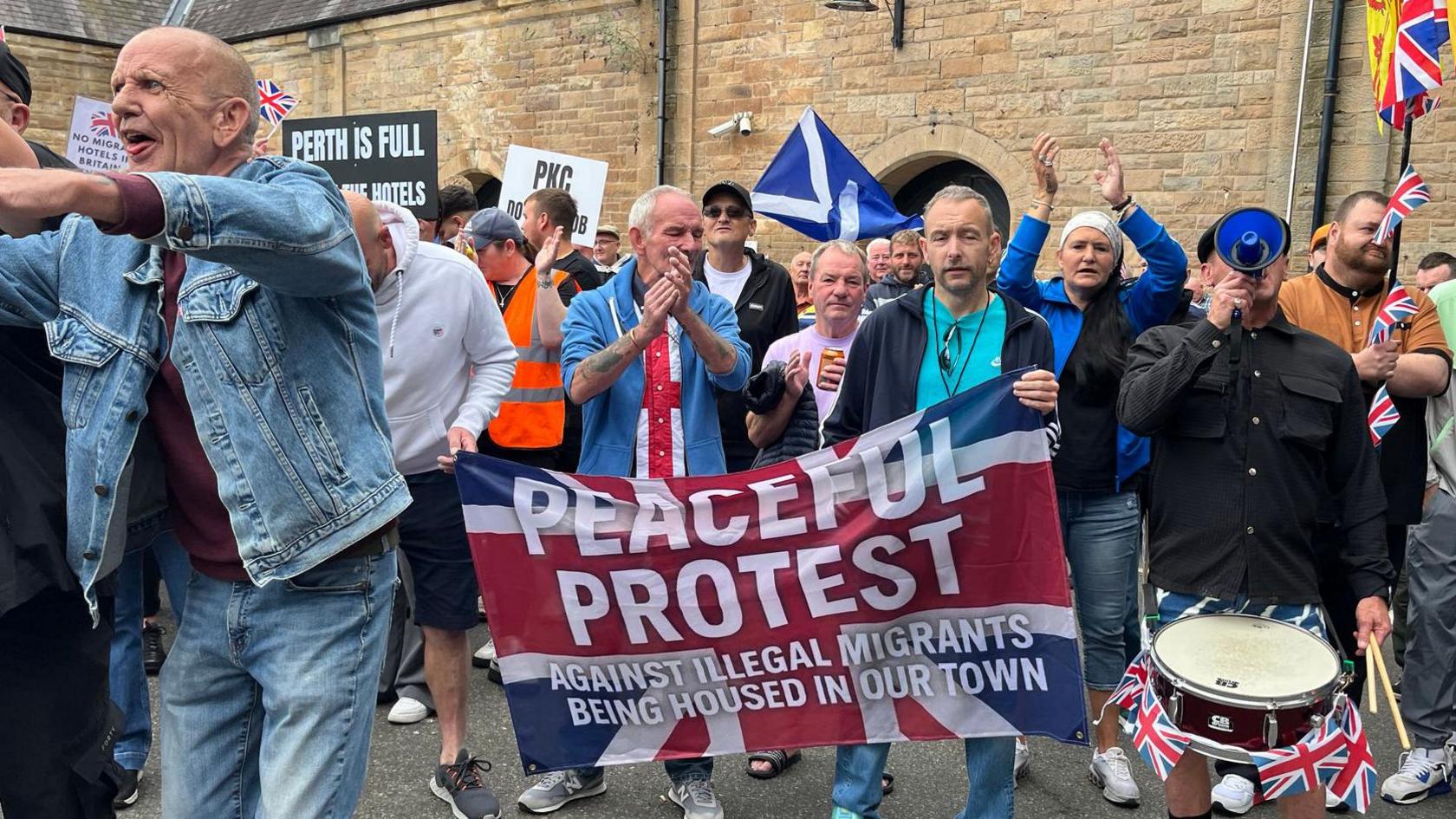 A group of people clapping and holding signs which say things like "perth is full" and "peaceful protest against illegal migrants being housed in our town"