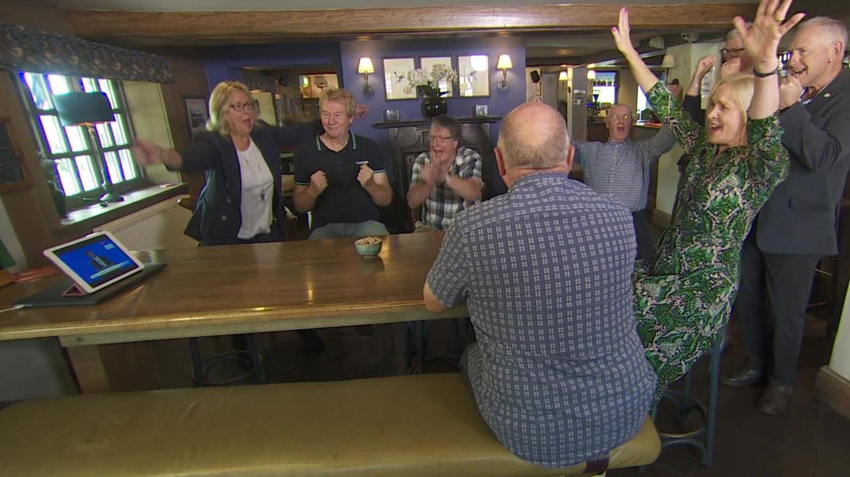 People gather around a tablet computer on a large table in a country pub. They are listening to the government announcement of the northern leg of HS2 being scrapped.