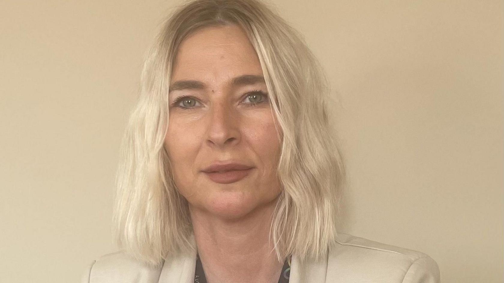 Mae gan Tricia Roberts wallt melyn hyd at ei hysgwyddau ac mae hi'n sefyll yn erbyn cefndir hufen. Mae hi'n gwisgo siaced hufen ac mae ganddi fynegiant niwtral ar ei hwyneb wrth iddi edrych ar y camera.